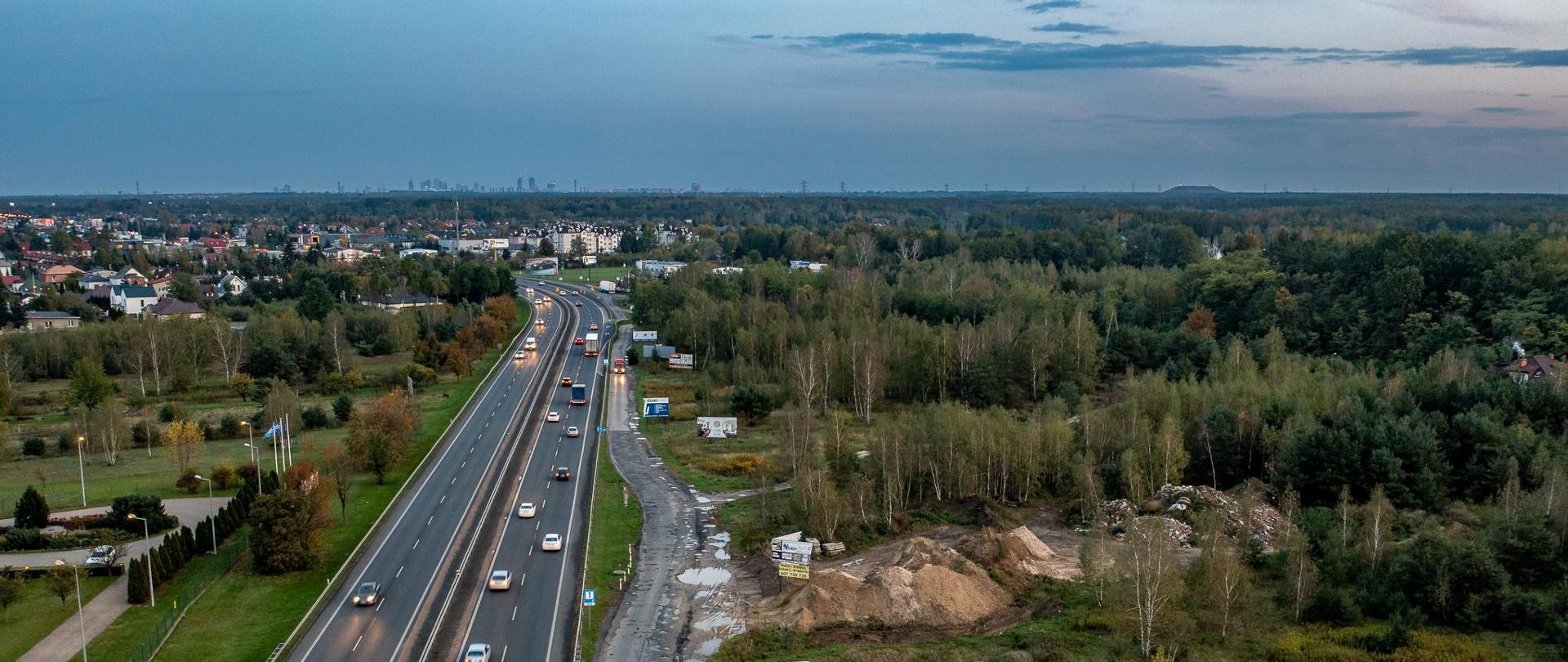 Zdjęcie przedstawia widok z lotu ptaka na drogę o wielu pasach ruchu, na której panuje umiarkowany ruch. Po obu stronach drogi znajdują się obszary z zielenią, drzewami, a po prawej stronie widoczna jest także rozpoczęta budowa. W oddali widać panoramę miasta na tle horyzontu. Niebo jest zachmurzone.