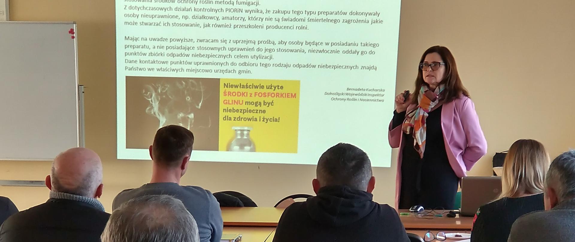 Siedzący w ławkach uczestnicy szkolenia patrzący w kierunku wyświetlonej na ścianie prezentacji. Obok w fioletowym żakiecie stoi przedstawicielka Oddziału w Strzelnie.