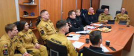 Strażacy uczestniczą w szkoleniu, będąc w biurze komendy wojewódzkiej PSP, wpatrując się w ekran telewizora, na którym prezentowana jest treść szkolenia.