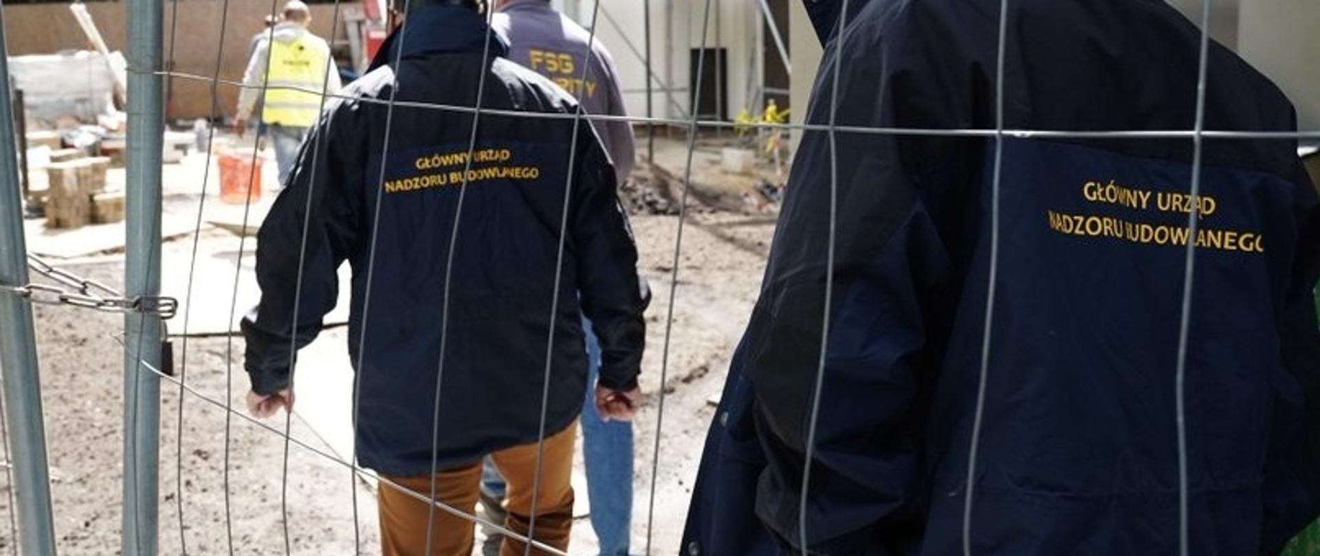 „Grupa osób w kurtkach z napisem GUNB na placu budowy, za ogrodzeniem, w tle sprzęt i materiały budowlane.”