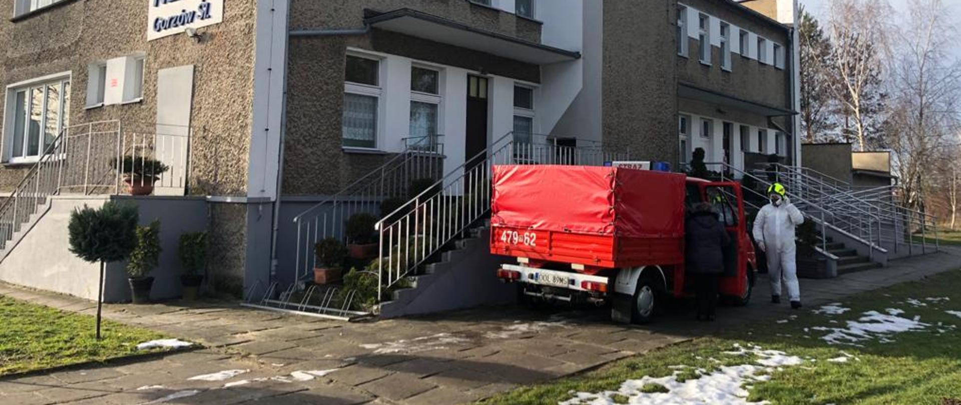 Na zdjęciu widoczny budynek przychodni przed którym stoi samochód jednostki OSP Gorzów Śląski - kolor czerwony. Przy pojeździe strażak OSP ubrany w kombinezon ochronny koloru białego i hełm koloru kanarkowego.