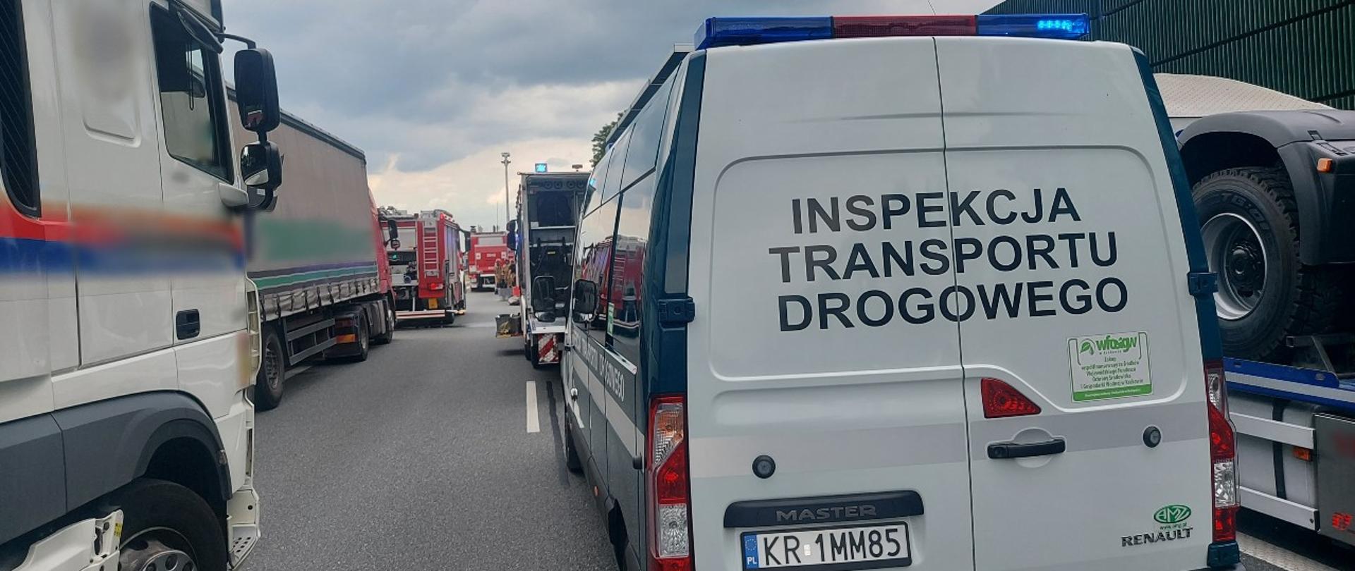 Na pierwszym planie tył oznakowanego radiowozu typu furgon małopolskiej ITD i ciężarówki stojące w korku przed miejscem wypadku. W tle stoją samochody ratowniczo-gaśnicze Straży Pożarnej. 