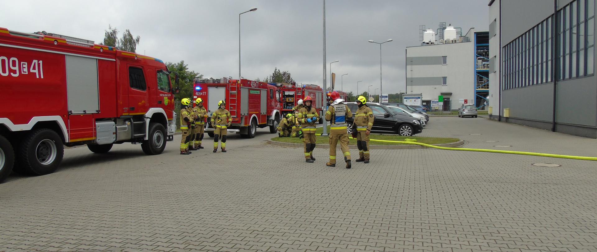 Zdjęcie przedstawia strażaków biorących udział w ćwiczeniach, po prawej budynki zakładu a po lewej samochody straży pożarnej w tle pochmurne niebo