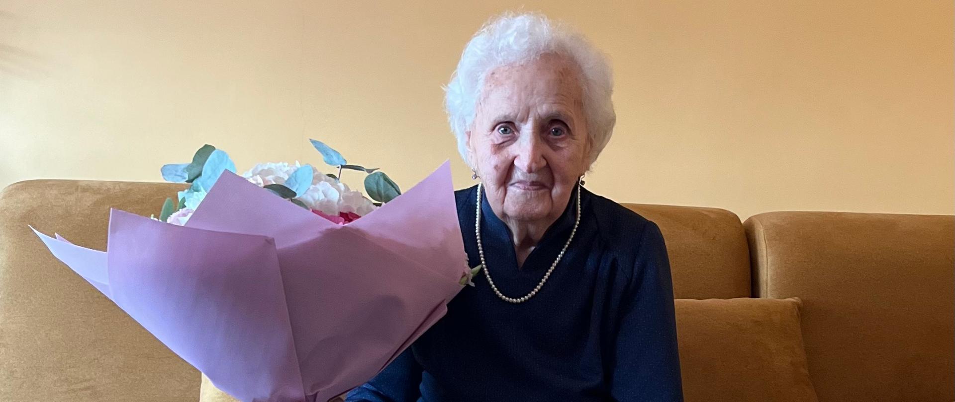 Na kanapie siedzi Jubilatka trzyma w ręku bukiet pięknych kwiatów. Jest przejęta podniosłą chwilą i delikatnie się uśmiecha.