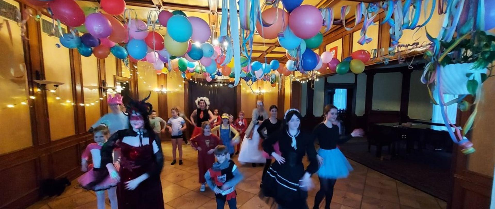 Zdjęcie kolorowe. Grupa osób w różnorodnych kostiumach karnawałowych tańcząca w udekorowanej sali. Pod sufitem wiszą liczne kolorowe balony (niebieskie, czerwone, żółte, różowe) oraz wielobarwne serpentyny. Ściany pomieszczenia wyłożone są drewnianą boazerią, a na podłodze ułożone są jasne płytki z ozdobnym wzorem.
W pierwszym planie kobieta w długiej, bordowej sukni i ciemnym nakryciu głowy z rogami oraz chłopiec w stroju rycerza. Obok nich postać w czarnej sukience z białym kołnierzykiem i charakterystycznymi warkoczami oraz dziewczyna w niebieskiej spódniczce tiulowej. W tle pozostali uczestnicy zabawy, w tym dzieci w kostiumach, m.in. w stroju minionka oraz różowym przebraniu. Sala jest oświetlona ciepłym światłem lampek ściennych i girland świetlnych.