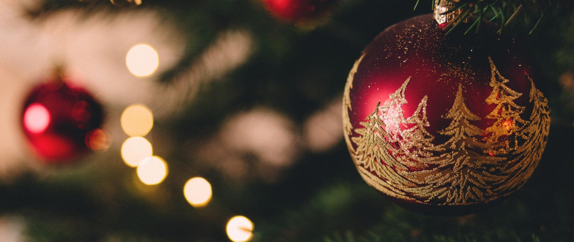 Zdjęcie przedstawia czerwona bombkę z złotymi choinkami. W tle widać kolejna bombkę oraz rozmytą choinkę.
