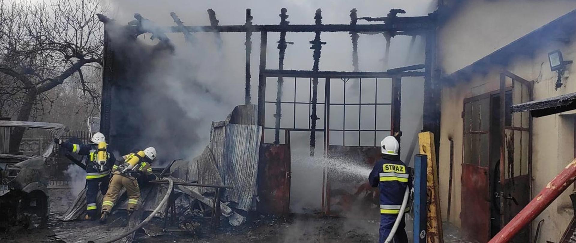 Zdjęcie zrobione przed spalonym budynkiem lakierni. Działanie gaśnicze prowadzi trzech strażaków podających dwa prądy gaśnicze wody do wnętrza budynku. Z budynku lakierni, przez spalony dach wydobywają się jasne kłęby dymu.