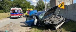 Na pierwszym planie rozbity samochód na poboczu drogi. W tle dwie karetki pogotowia ratunkowego. 