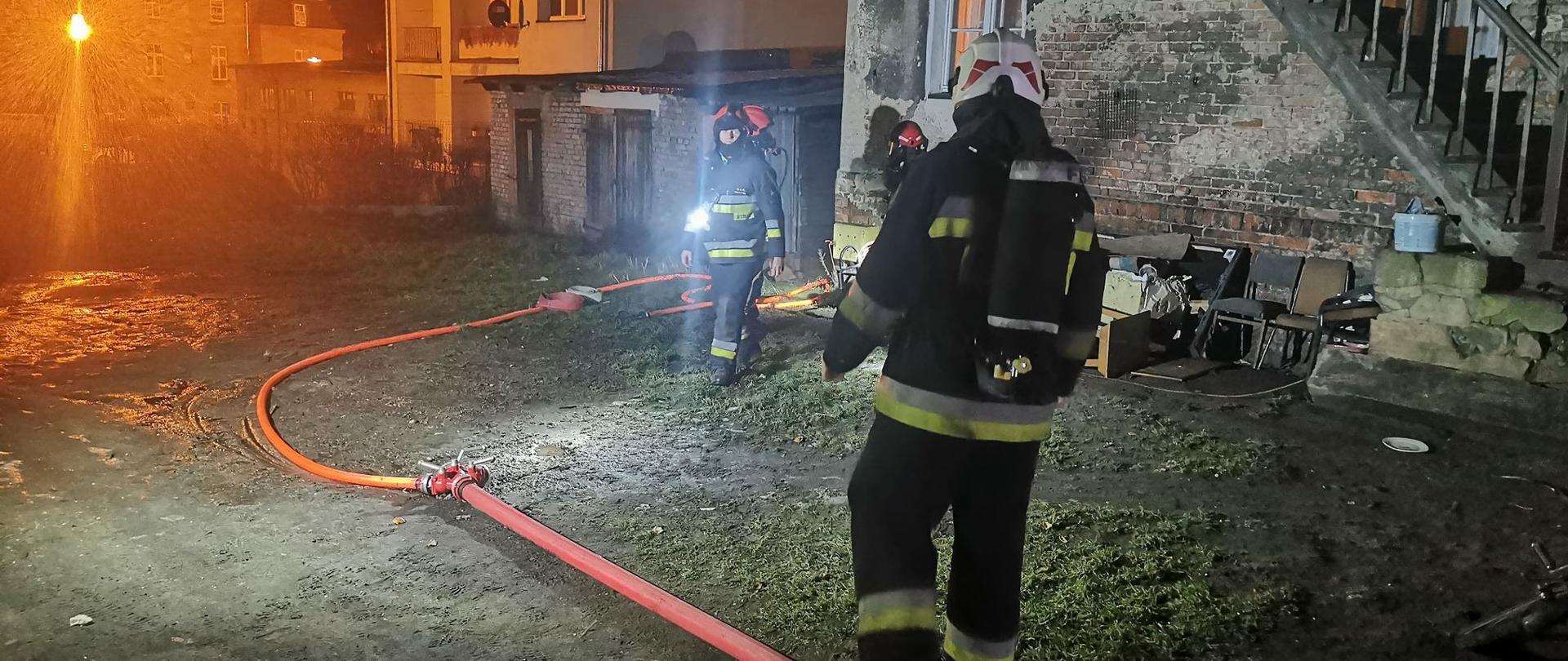 Zdjęcie przedstawia dwóch strażaków ubranych w ubranie specjalne, hełmy, sprzęt ochrony dróg oddechowych. W centrum zdjęcia widać grubszy czerwony wąż strażacki zakończony rozdzielaczem do którego zaś podłączony jest do lewej nasady kolejny cięższy czerwony wąż strażacki. W tle zdjęcia widać stary budynek do którego przylegają schody prowadzące na piętro. W tle również widać bloki oraz mocno świecącą na pomarańczowo lampę uliczną. Zdjęcie zrobione nocą.