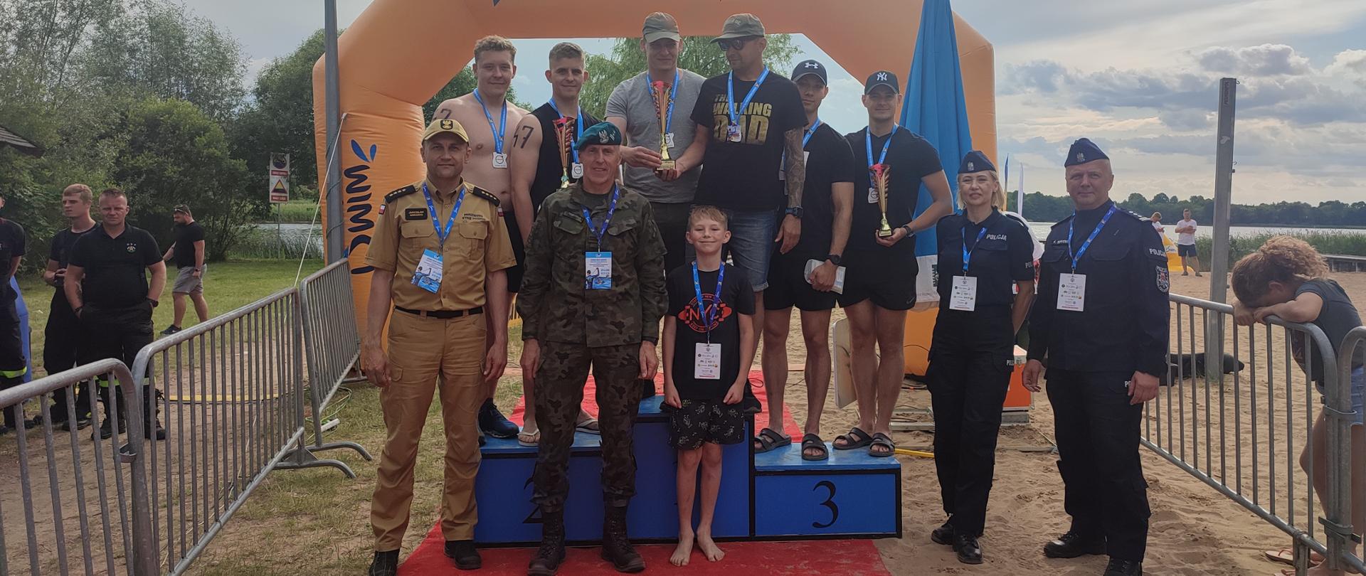 Mężczyźni stoją na podium. Przy nich funkcjonariusze straży pożarnej, policji oraz wojska