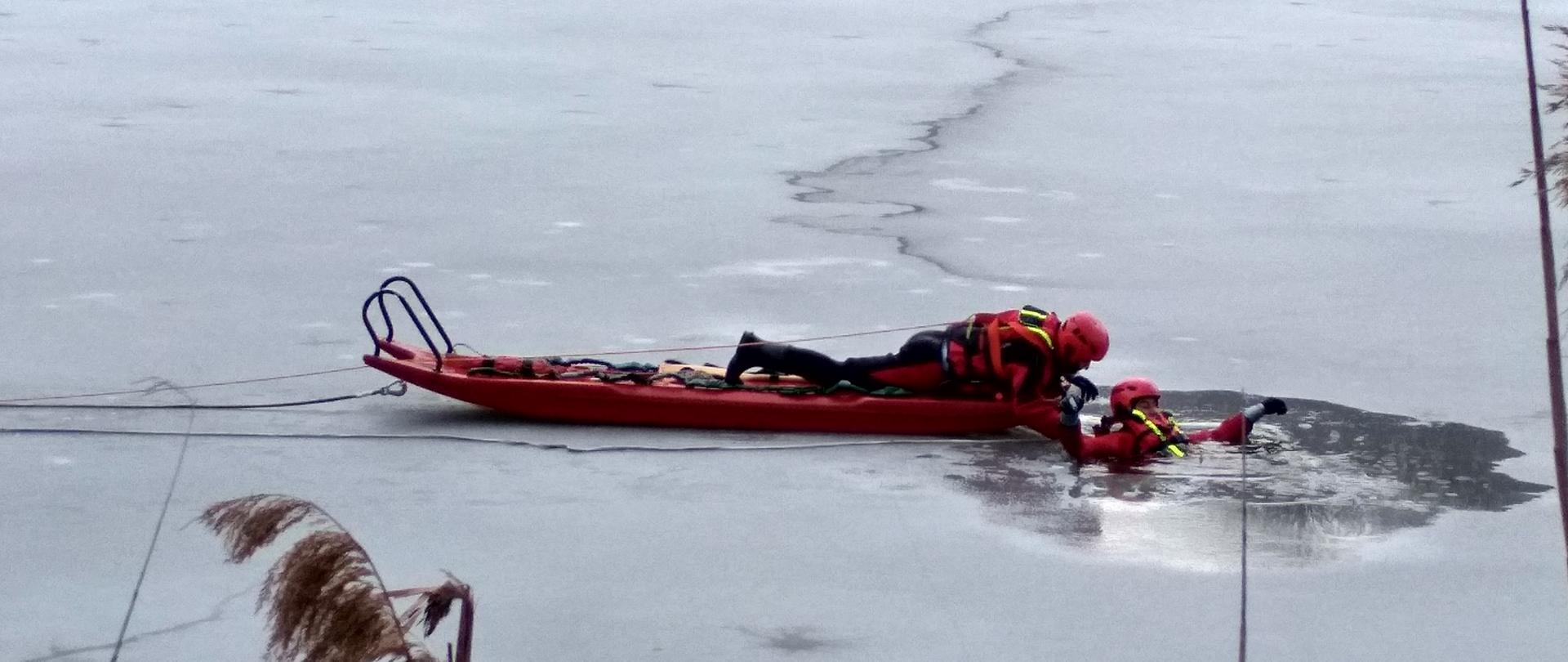 Na zdjęciu widoczny zamarznięty akwen, oraz strażacy podczas ćwiczeń w podejmowaniu człowieka pod którym załamał się lód.