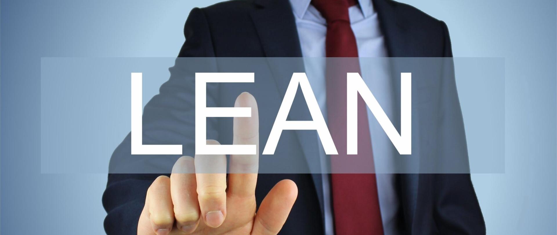 Na zdjęciu widać ubranego w garnitur mężczyznę. Nie widać jego głowy, widać tylko korpus. Jego ręka, palcem wskazującym, dotyka tabliczki z napisem LEAN.
