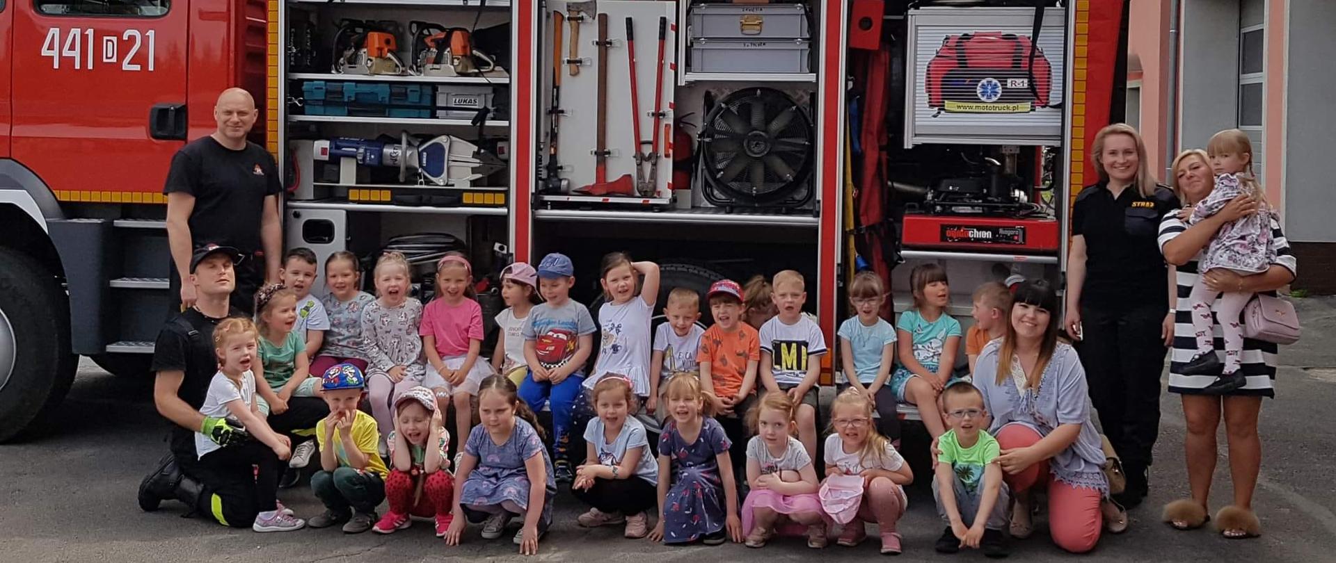 Obraz przedstawia grupę przedszkolną i strażaków w trakcie wizyty w jaworskiej komendzie. Osoby pozują do wspólnego zdjęcia na tle samochodu pożarniczego.