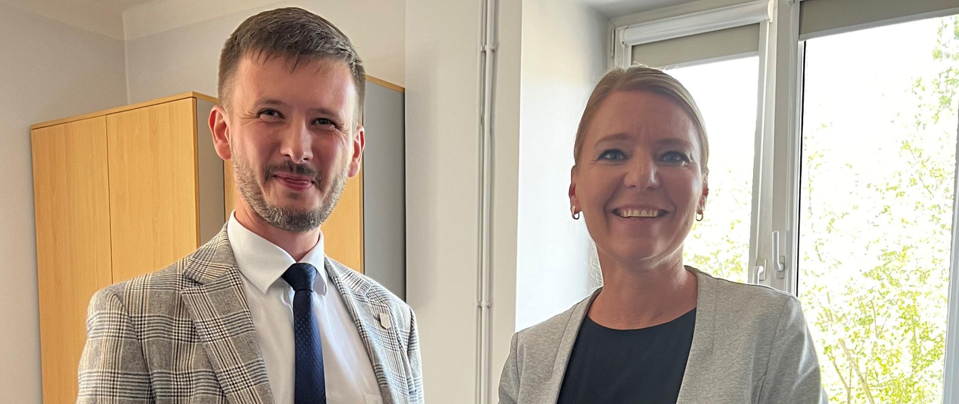 Dwie osoby stojące w pomieszczeniu; jedna w jasnej marynarce w kratę, druga w szarej marynarce, w tle szafka i okna.