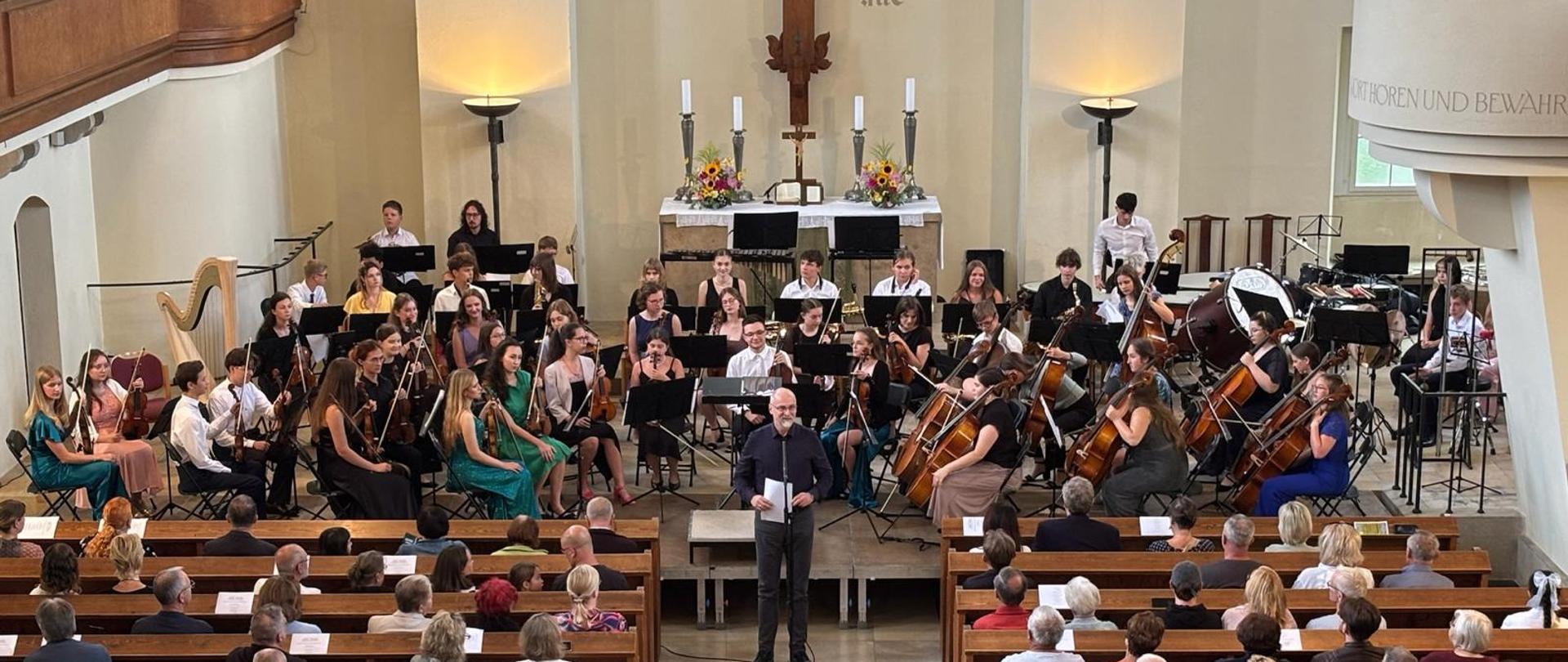 Orkiestra symfoniczna w kościele podczas występu, publiczność w ławkach.