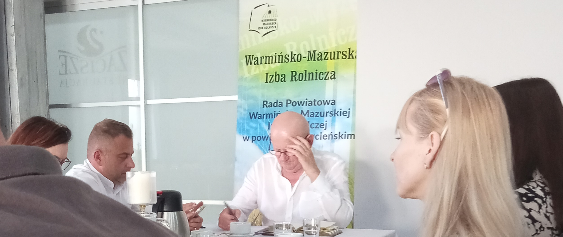 Wnętrze jasnej sali z białym wykończeniem, w tle baner Rady Powiatowej Warmińsko-Mazurskiej Izby Rolniczej w powiecie szczycieńskim. Na tle baneru za stołem siedzi mężczyzna w okularach podpierający głowę. Stół nakryty białym obrusem, na stole szklanki z napojami oraz dokumenty. Wokół stołu siedzą 3 kobiety i 2 mężczyzn.