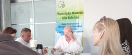 Wnętrze jasnej sali z białym wykończeniem, w tle baner Rady Powiatowej Warmińsko-Mazurskiej Izby Rolniczej w powiecie szczycieńskim. Na tle baneru za stołem siedzi mężczyzna w okularach podpierający głowę. Stół nakryty białym obrusem, na stole szklanki z napojami oraz dokumenty. Wokół stołu siedzą 3 kobiety i 2 mężczyzn.