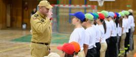 Grupa młodych ludzi w jasnych koszulkach i kolorowych czapkach stoi w rzędzie na sali gimnastycznej. Z lewej strony, w piaskowym mundurze, stoi strażak, salutując w kierunku stojących przed nim osób.