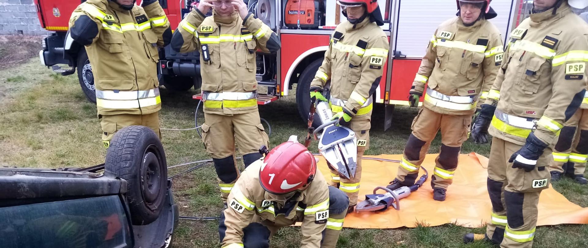 Zdjęcie wykonane w ciągu dnia. Widać wrak pojazdu osobowego. Przy pojeździe znajdują się funkcjonariusze Państwowej Straży Pożarnej w umundurowaniu bojowym, w kolorze piaskowym z czerwonymi hełmami. Posiadają sprzęt ratowniczy. Zdjęcie wykonane na podłożu trawiastym.