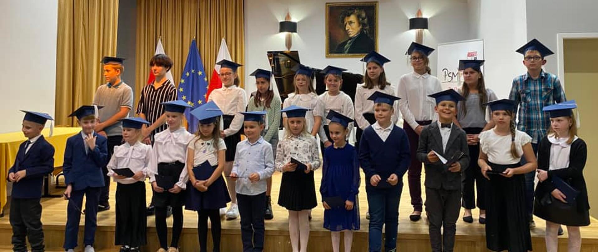 Zdjęcie przedstawiające wszystkich uczniów klas pierwszych podczas pasowania na ucznia Państwowej Szkoły Muzycznej I st. im. Fryderyka Chopina w Miastku. Uczniowie ustawieni w dwóch rzędach na scenie salki koncertowej szkoły.