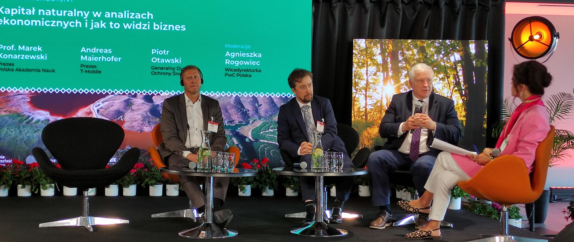 Na scenie siedzi 3 mężczyzn i kobieta. Jeden z mężczyzn trzyma mikrofon. Za ich plecami na ścianie wyświetlony slajd z nazwiskami prelegentów.