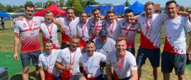 Grupa polskich strażaków w sportowych strojach biało - czerwonych w dwóch rzędach pozuje do zdjęcia. Na szyjach mają zawieszone medale