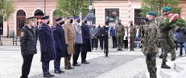 Na zdjęciu po prawej stronie żołnierz w mundurze maskującym podczas pełnienia warty honorowej. Drugi, tak samo ubrany żołnierz wchodzi po kamiennych schodach z wieńcem kwiatów. Po lewej stronie sześciu funkcjonariuszy w mundurach galowych stoi w szeregu. Wszyscy mają założone maseczki ochronne. W tle kolorowe fasady kamienic i pozostali uczestnicy uroczystości.
