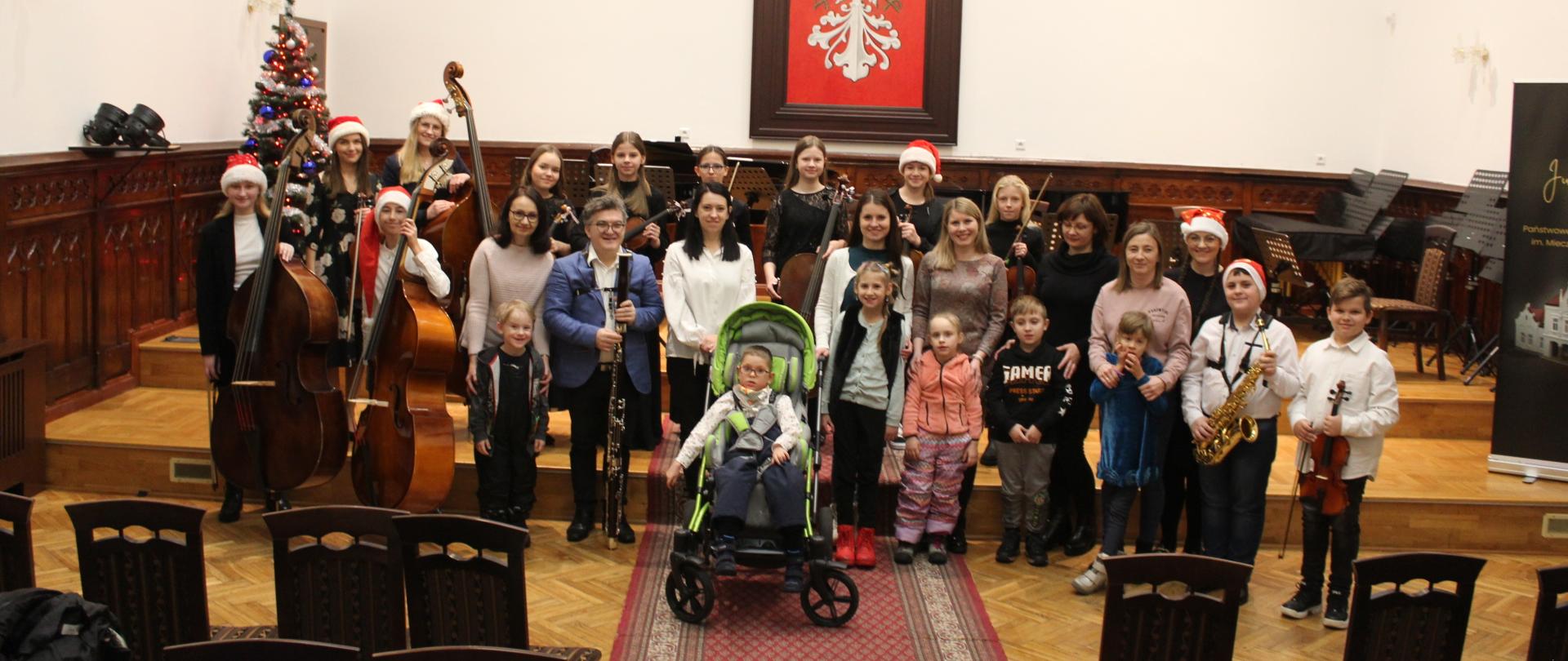 zdjęcie przedstawia grupę wychowanków Powiatowego Zespołu Placówek Szkolno-Wychowawczych w Mielcu oraz uczniów naszej szkoły z klasy kontrabasu, saksofonu, skrzypiec i fortepianu, na scenie w sali królewskiej Państwowej Szkoły Muzycznej w Mielcu, w tle widać godło Polski