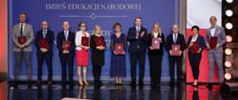 Pod niebieską ścianą z napisem Dzień Edukacji Narodowej stoi w szeregu grupa kilkunastu osób, trzymają czerwone teczki.