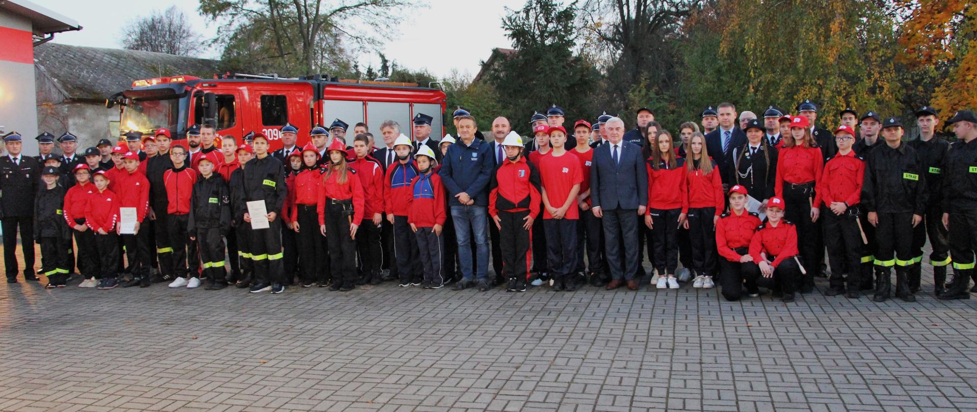 Zdjęcie przedstawia zaproszonych gości i organizatorów wśród przedstawicieli Młodzieżowych Drużyn Pożarniczych i ich opiekunów oraz prezesów ochotniczych straży pożarnych na pamiątkowym ustawieniu podczas uroczystości.