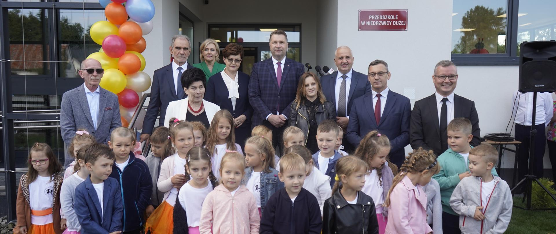 Na pierwszym planie grupa dzieci, za nimi grupa dorosłych. Wszyscy pozują do zdjęcia przed wejściem do przedszkola. 