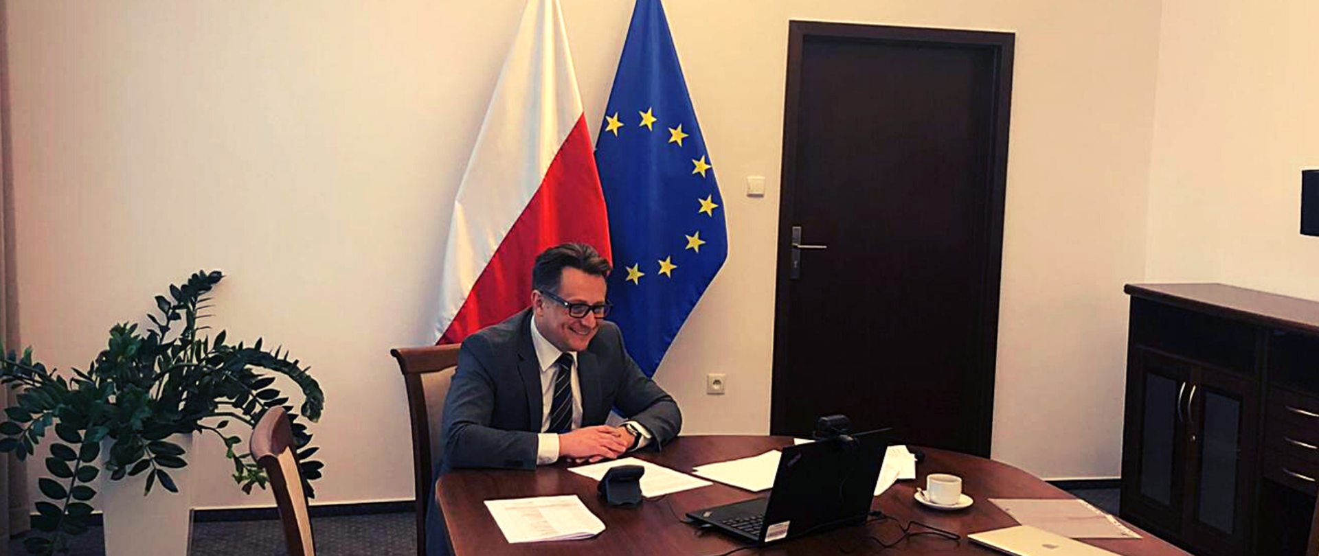 Minister Krzysztof Szubert podczas spotkania online - siedzi przed laptopem. Za nim flagi Polski i Unii Europejskiej.