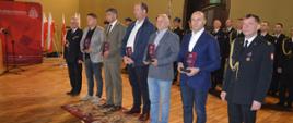 Dzień Strażaka w Rawiczu. Sala widowiskowa w domu kultury. Na tle pododdziału w szeregu stoją: z-ca komendanta powiatowego PSP, przedstawiciele firm i instytucji, którzy otrzymali okolicznościowe statuetki w podziękowaniu za wsparcie komendy, i komendant powiatowy PSP.