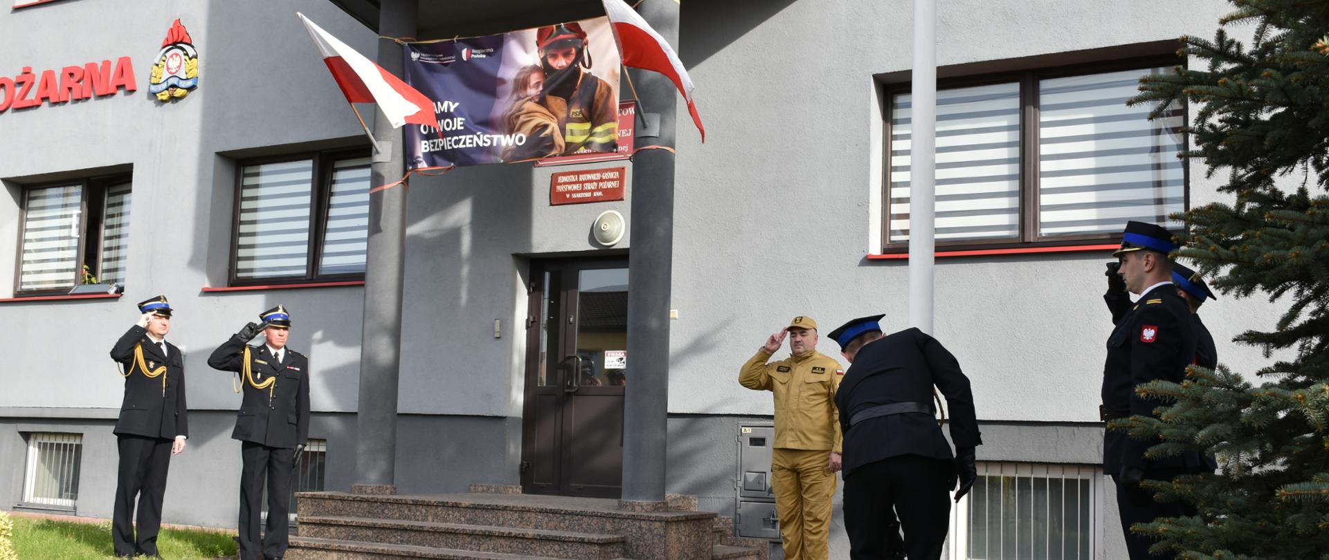 Zdjęcie obrazuje strażaków znajdujących się w poczcie flagowym, którzy zapieli flagę na masz. Wciągają oni flagę przy akompaniamencie hymnu Rzeczypospolitej Polskiej. 