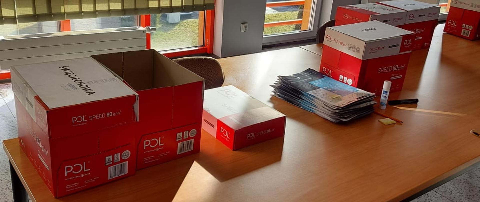 Kartony z ulotkami stoją na stole. Na wierzchniej części widoczne napisy miejscowości.