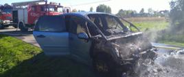 Na zdjęciu widać samochód osobowy ze spaloną komorą silnika, w oddali pojazdy gaśnicze OSP i PSP.