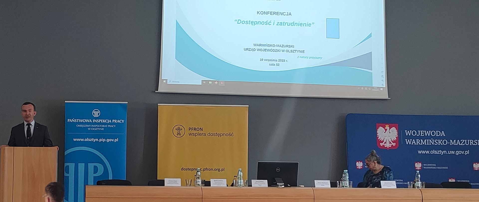 mężczyzna przemawia z mównicy, po prawej stół a nad nim ekran z prezentacją