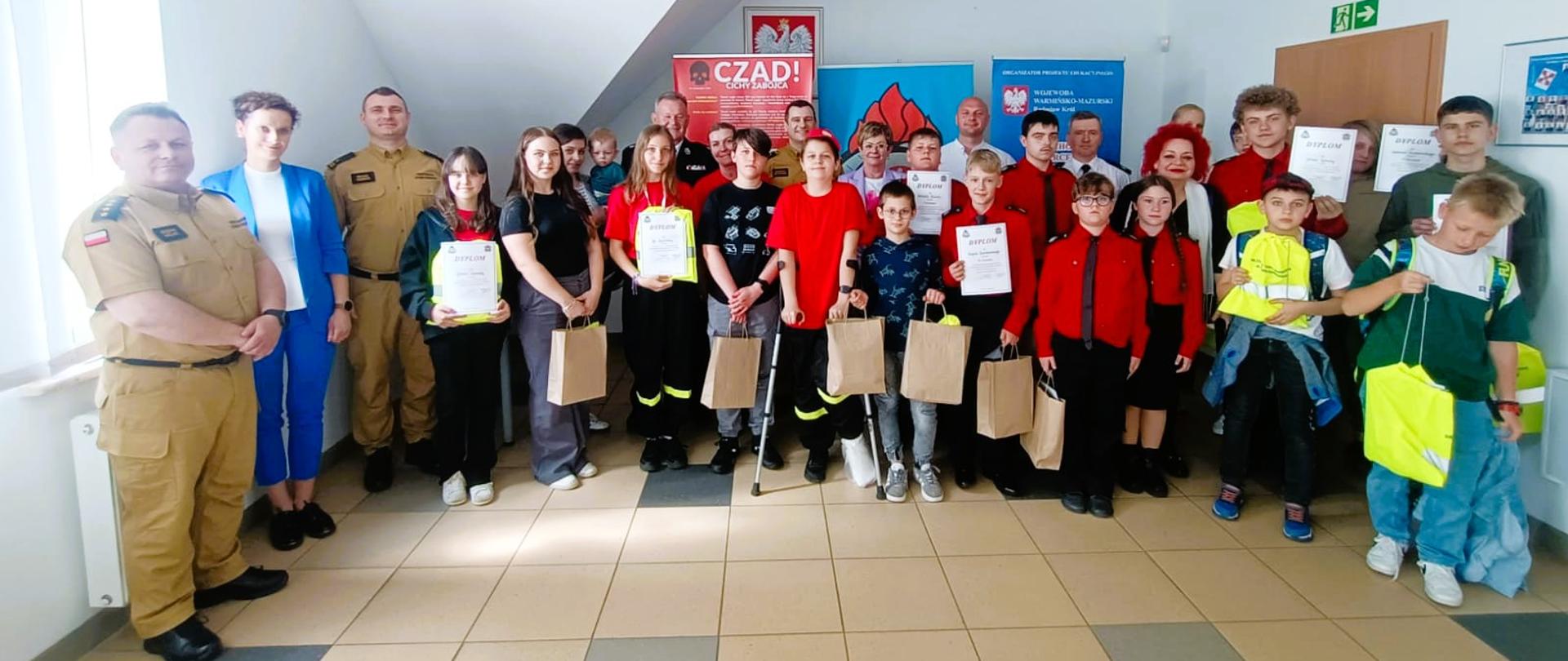 Zdjęcie przedstawia wszystkich uczestników eliminacji powiatowych Ogólnopolskiego Turnieju Wiedzy Pożarniczej „Młodzież Zapobiega Pożarom” z nagrodami/dyplomami oraz opiekunów z poszczególnych szkół i organizatorów przedsięwzięcia, zlokalizowanych w świetlicy Komendy Powiatowej Państwowej Straży Pożarnej w Gołdapi
