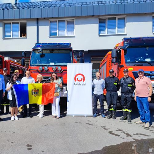 Grupa kobiet i mężczyzn, część w strojach strażackich, pozuje na tle wozów strażackich, z flagą Mołdawii i banerem z logo Polskiej pomocy