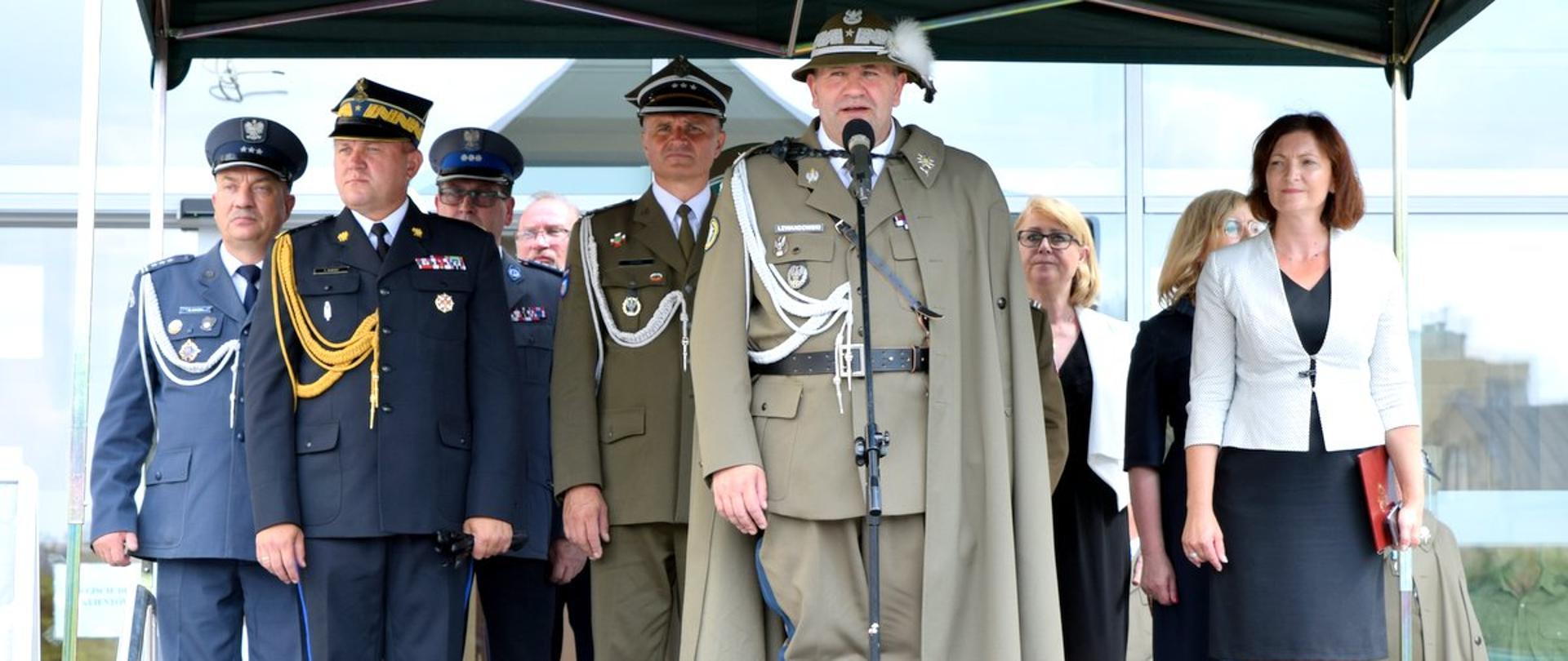 Przedstawiciele służb mundurowych stoją na podium. Od lewej strony przedstawiciele: Służby Więziennej, Państwowej Straży Pożarnej, Policji i Wojska Polskiego. Z przodu, przed mikrofonem stoi żołnierz ubrany w strój podhalańczyka w stopniu generała. Po prawej stronie 3 osoby w cywilu. 