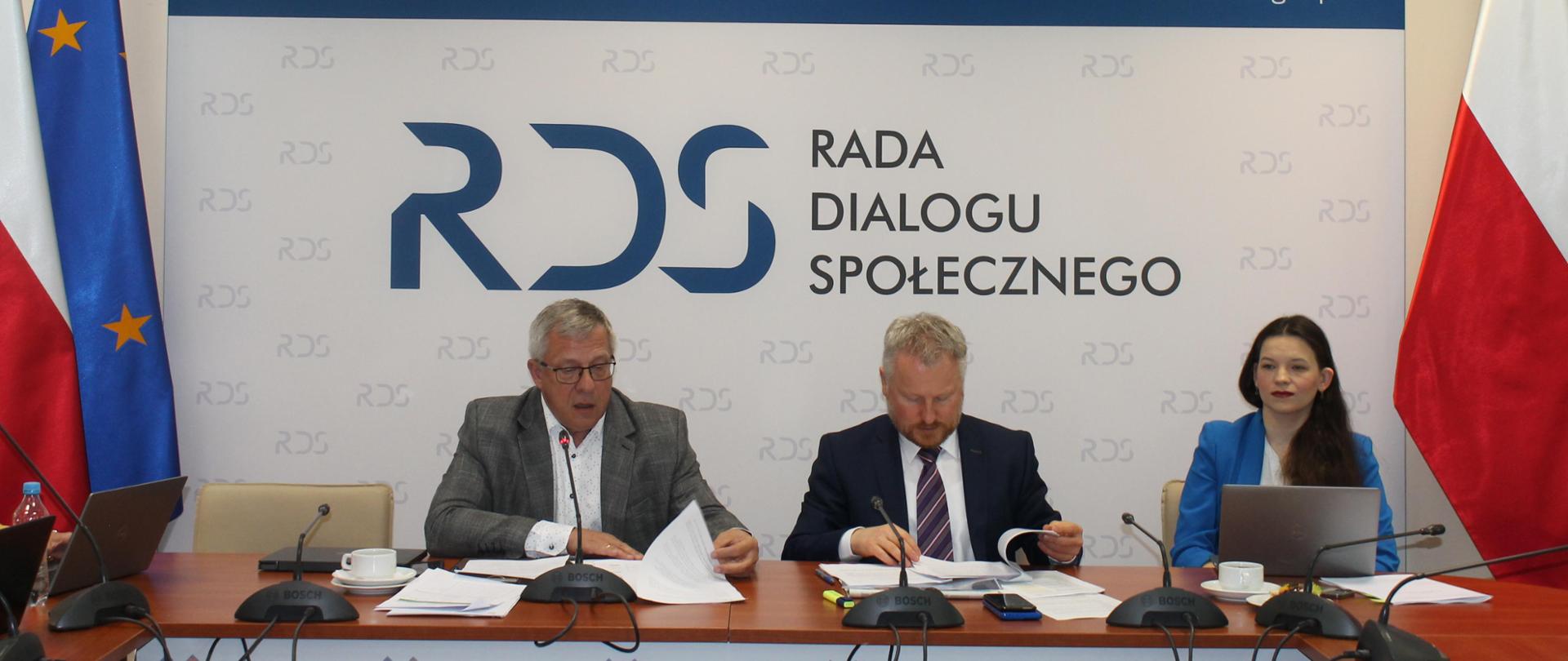 Na zdjęciu widnieje stół prezydialny za którym siedzą od lewej strony: Przewodniczący zespołu ds. budżetu, wynagrodzeń i świadczeń socjalnych – Leszek Miętek, Jurand Drop – Podsekretarz Stanu w Ministerstwie Finansów oraz Katarzyna Nowakowska - Podsekretarz Stanu w Ministerstwie Rodziny, Pracy i Polityki Społecznej. Po obu stronach stołu widnieją dwie flagi: Polski i UE. W tle siedzących znajduje się baner z napisem Rada Dialogu Społecznego a nad nim wisi godło Polski.