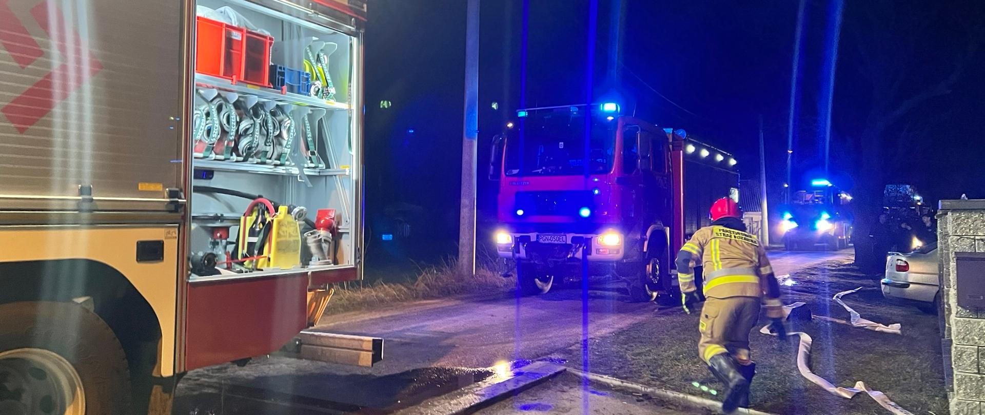 Na zdjęciu widać trzy samochody gaśnicze. Jeden ze4 strażaków rozwija linię wężową. Jest pora nocna, samochody pożarnicze oświetlają teren działań.