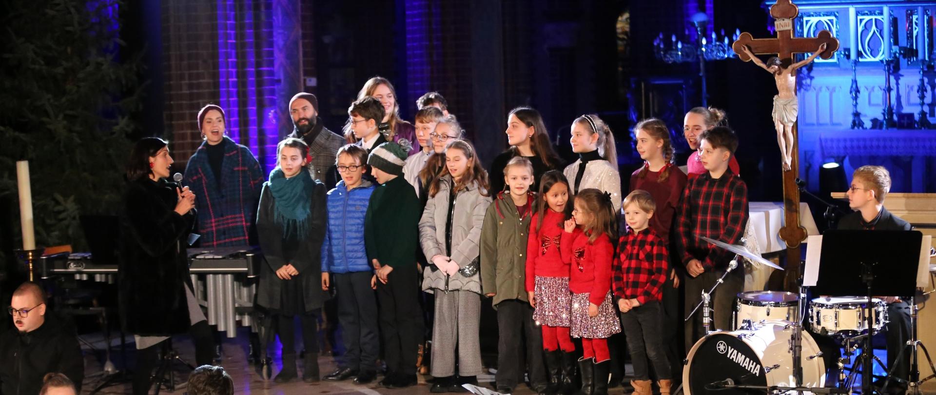 Na zdjęciu widać grupę dzieci stojących na podwyższeniu w bazylice św. Antoniego, podczas koncertu. Dzieci są ubrane ciepło i odświętnie — niektóre mają czerwone lub zielone ubrania, w związku z bożonarodzeniowym charakterem wydarzenia.
Po lewej stronie stoi kobieta z mikrofonem, która śpiewa z dziećmi. W tle widać ołtarz z krucyfiksem, a całość jest oświetlona kolorowym światłem — głównie niebieskim i fioletowym.
Na pierwszym planie, poniżej sceny, znajduje się orkiestra — muzycy grają na różnych instrumentach, m.in. skrzypcach, wiolonczeli, fagocie i perkusji. Cała scena ma uroczysty, koncertowy nastrój.