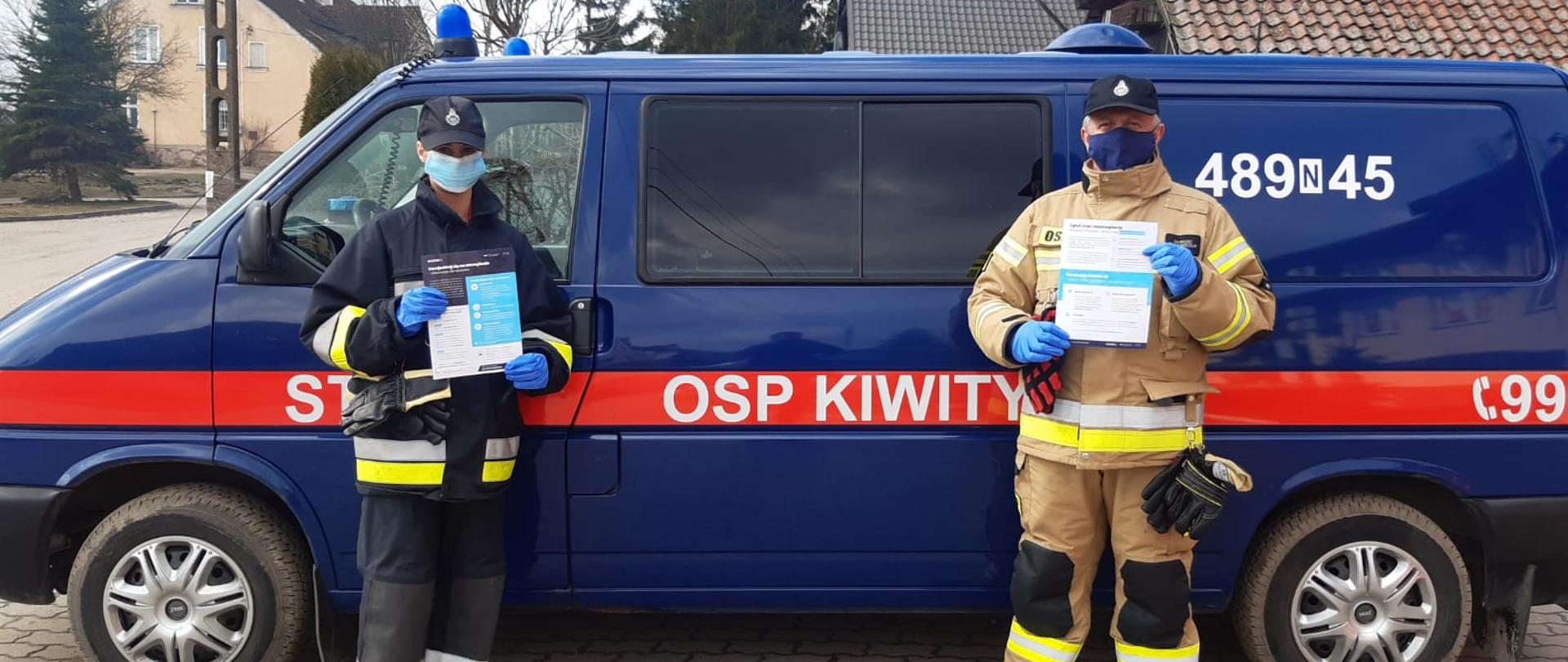 Na zdjęciu dwóch strażaków OSP na tle samochód. W dłoniach maja ulotki dot. akcji szczepień