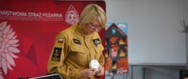 Na zdjęciu widoczna jest kobieta w mundurze strażackim, stojąca przy stole podczas prezentacji lub szkolenia. Trzyma w dłoniach czujnik dymu i przygląda się mu z uwagą. Na stole przed nią znajduje się otwarta walizka z wyposażeniem, m.in. gaśnicą i innymi akcesoriami związanymi z bezpieczeństwem pożarowym. W tle widać czerwony baner Państwowej Straży Pożarnej oraz planszę edukacyjną przedstawiającą zagrożenia pożarowe w domu. Atmosfera sugeruje pokaz lub instruktaż dotyczący ochrony przeciwpożarowej.