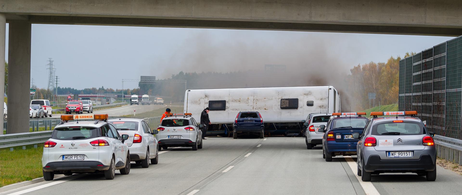 Na autostradzie pod wiaduktem stoi przewrócona przyczepa kempingowa blokująca oba pasy ruchu. Wokół niej znajdują się samochody służb drogowych z włączonymi światłami ostrzegawczymi. Z przyczepy unosi się ciemny dym. Na wiadukcie nad drogą widoczni są ludzie obserwujący sytuację.