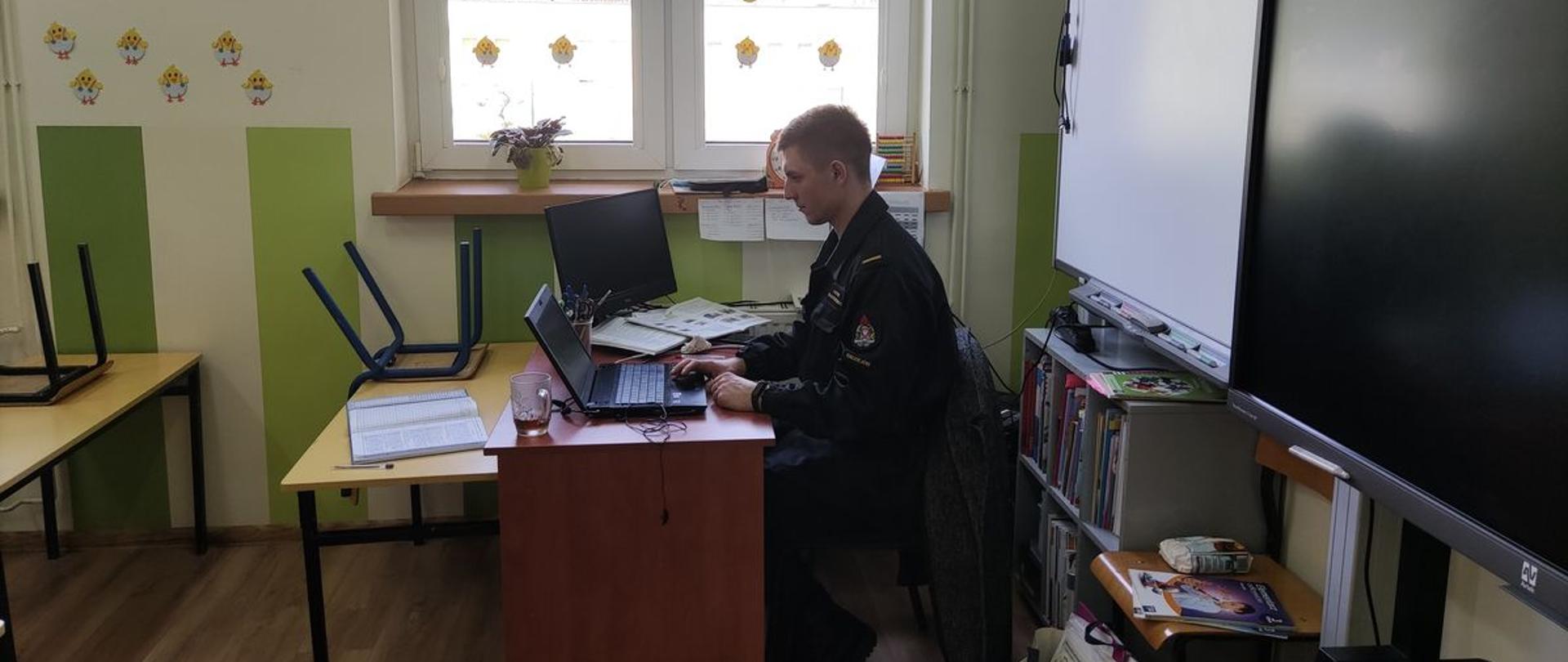 Zdjęcie przestawia funkcjonariusza Komendy Powiatowej Państwowej Straży Pożarnej
w Radziejowie, siedzącego przy biurku nauczyciela w jednej z klas Szkoły Podstawowej im. Stefana Kardynała Wyszyńskiego w Bieganowie, który korzystając z laptopa nauczyciela prowadzi zajęcia online z dziećmi klas I-III w ramach kampanii edukacyjno-informacyjnej „Czujka na straży Twojego bezpieczeństwa”.