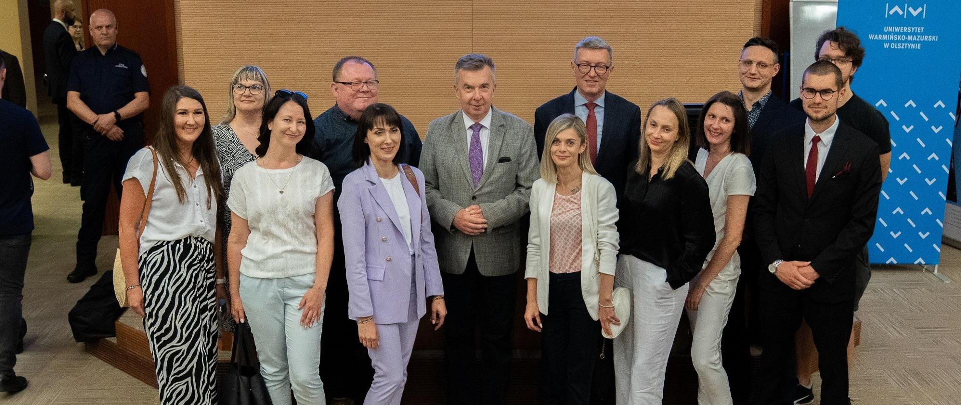 Zdjęcie zbiorowe, pod brązową ścianą stoi grupa kilkunastu osób, pośrodku minister Wieczorek.