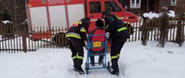 Na zdjęciu widać strażaków OSP w ubraniach koszarowych, którzy prowadzą inwalidzki wózek do samochodu ratowniczego. W oddali widać dom. Aura jest zimowa.