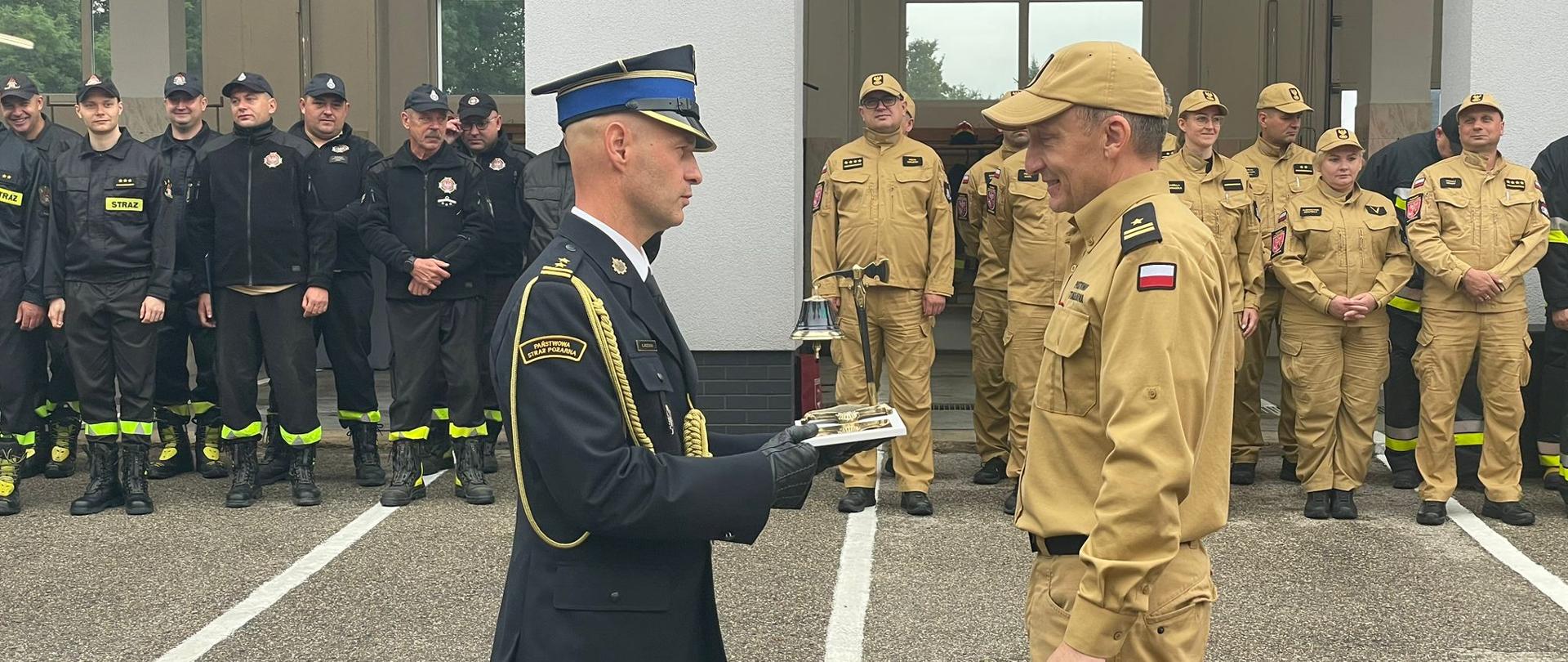 Komendant Miejski wręcza upominek nowemu emerytowi. W tle ustawieni w szeregu strażacy, dalej widoczny budynek remizy w Pasłęku.